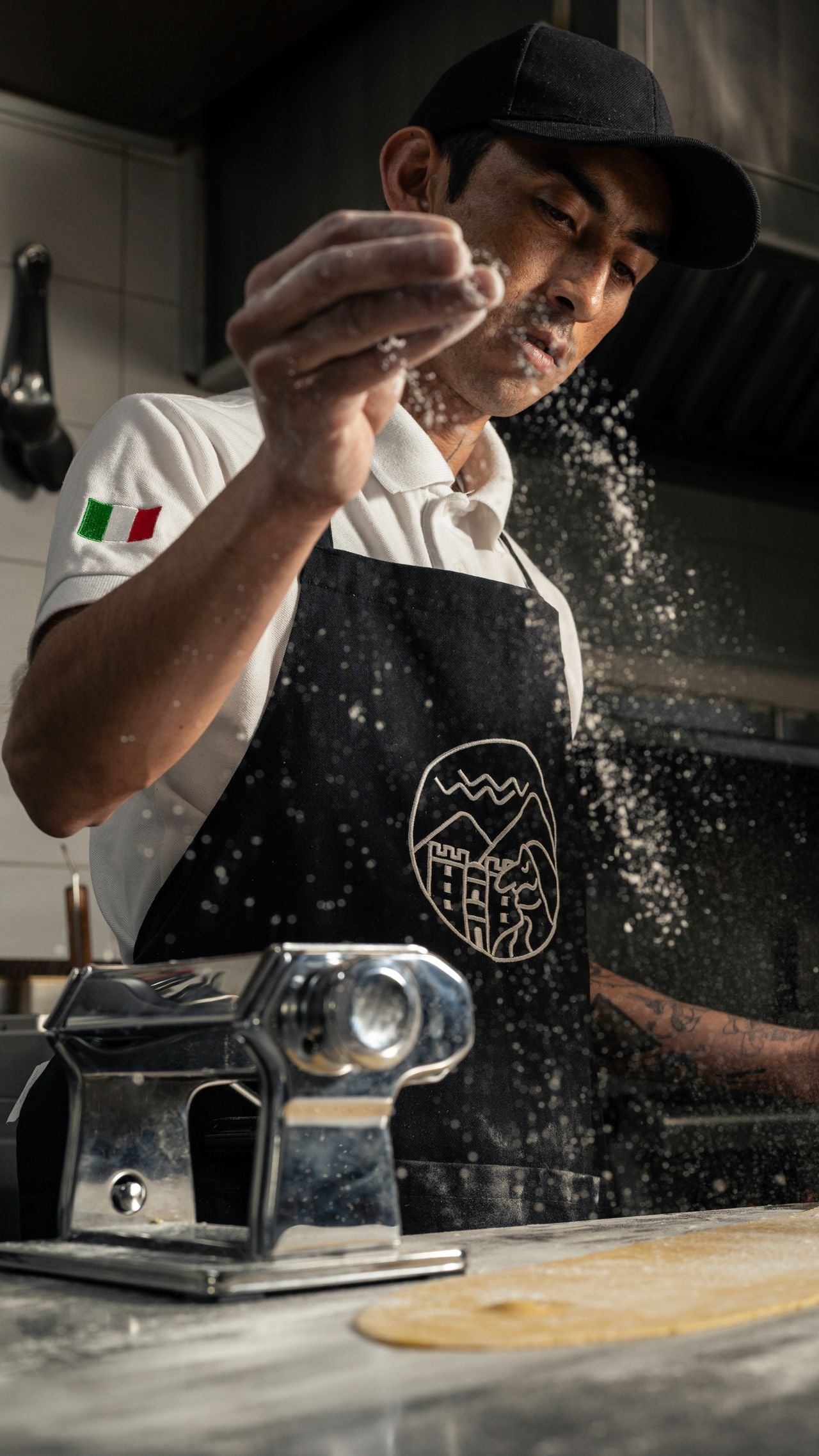 Person in a kitchen wearing a branded apron, working with dough and a pasta machine.